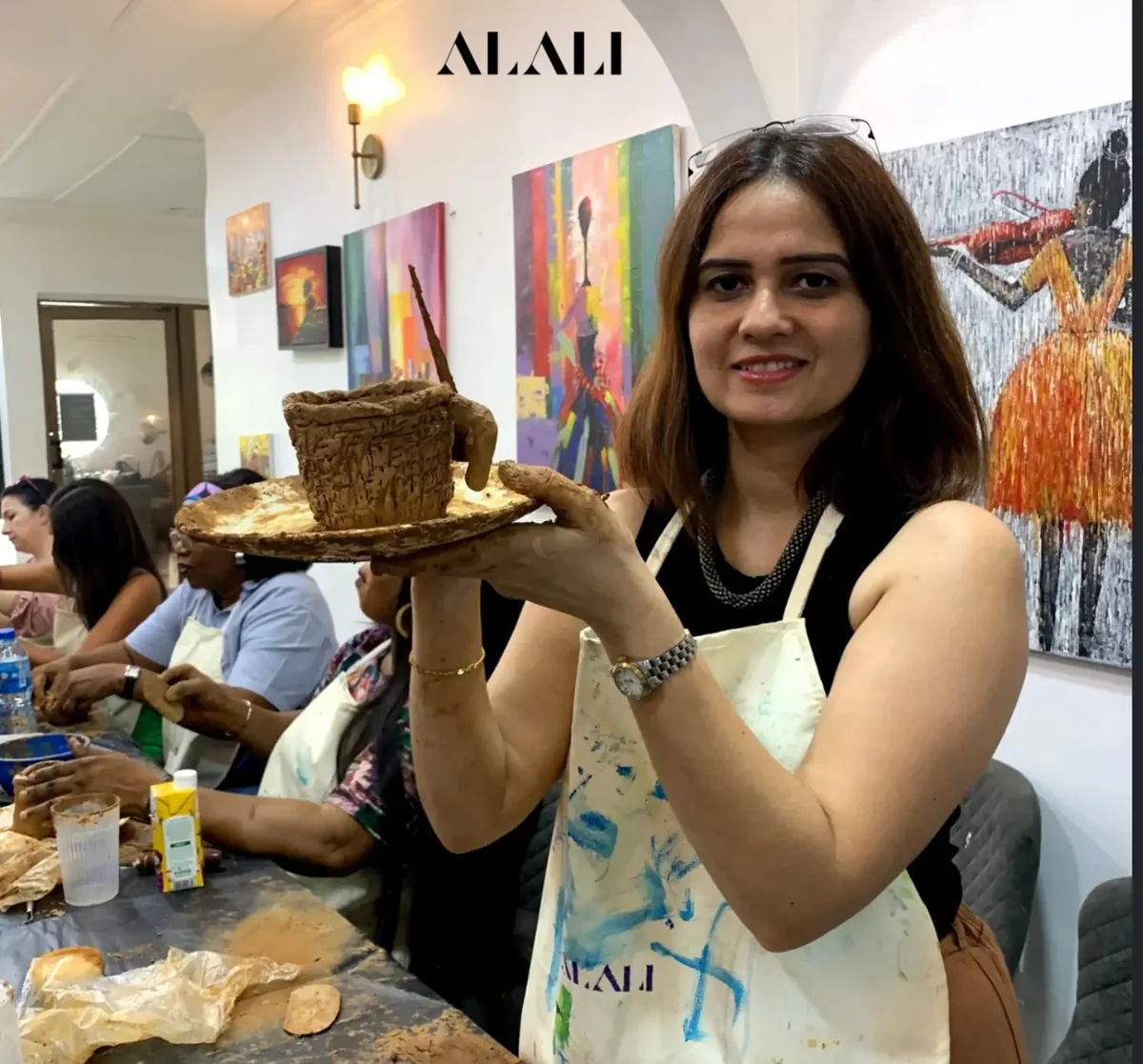A guest at Alali holding up her hand-built clay cup and plate, with other guests making pottery behind her